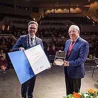 Zwei Männer im Anzug stehen auf einer Bühne, im Hintergrund ist das Publikum zu sehen. Die Männer halten eine Urkunde und eine Medaille.
