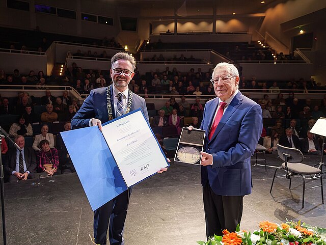 Zwei Männer im Anzug stehen auf einer Bühne, im Hintergrund ist das Publikum zu sehen. Die Männer halten eine Urkunde und eine Medaille.
