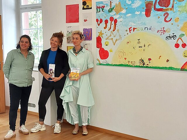 Drei Frauen stehen neben einem Bild, das von Kindern gemalt wurde. Auf dem Bild ist der Titel "Freunde" zu lesen.