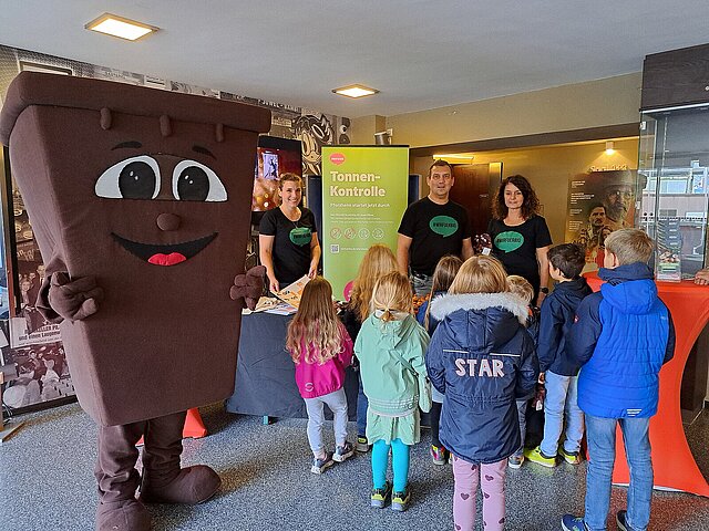 Eine Bio-Tonne als Maskottchen steht an einem Stand der Abfallwirtschaft. Davor stehen Kinder.