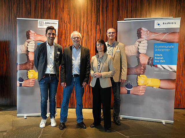 Auf dem Bild sind Christian Fuchs, stellvertretender Leiter des Jobcenters Enzkreis, Hartmut Schölch, Leiter des Jobcenters Enzkreis, Natalie Hohenstein, Leiterin des Pforzheimer Jobcenters und der stellvertretende Leiter des Jobcenters Pforzheim, Reiner Walter, zu sehen, die die Zeitarbeitsmesse im Reuchlinhaus eröffnen. Links und rechts von ihnen stehen zwei Banner der Jobcenter Enzkreis und Pforzheim.