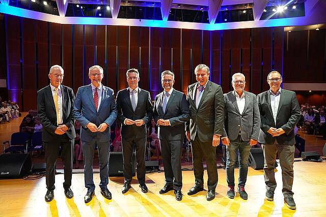 Sieben Männer in Anzügen auf einer Bühne