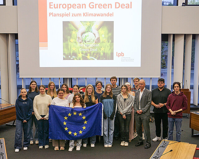 Schülerinnen und Schüler der Carlo Schmid Schule, Europabeauftrage Anna-Lena Beilschmidt und Bürgermeister Frank Fillbrunn stehen als Gruppe im Großen Sitzungssaal. Drei Schülerinnen halte eine EU-Flagge hoch und im Hinterrund ist eine Power-Point Präsentation der Landeszentrale für politische Bildung Baden-Württemberg auf der Leinwand zu sehen. Der Titel der Präsentation lautet "European Green Deal - Planspiel zum Klimawandel".