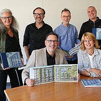 Sechs Personen stehen und sitzen an einem Tisch und präsentieren ein Buch