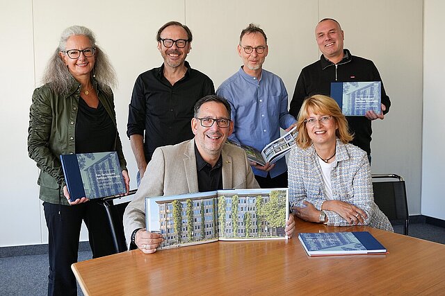 Sechs Personen stehen und sitzen an einem Tisch und präsentieren ein Buch