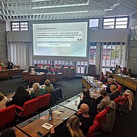 Viele Menschen sitzen in einem Sitzungssaal und schauen auf eine Präsentation