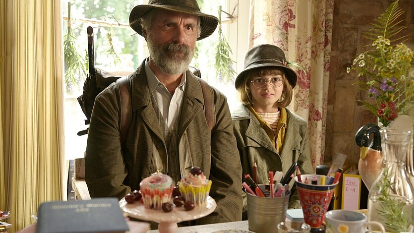 Der Buchspazierer Ein bärtiger Mann und ein Kind mit Brille stehen in einem gemütlichen Raum hinter einem Tisch mit Kuchen und Stiften. - copyright: KoKi - Ersteller: KoKi