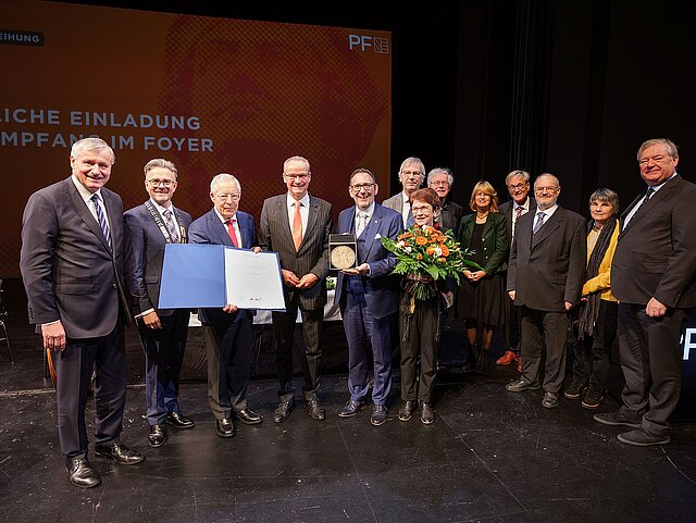Eine Gruppe von Menschen stehen im Halbkries und halten einen Blumenstrauß, eine Urkunde und eine Medaille.