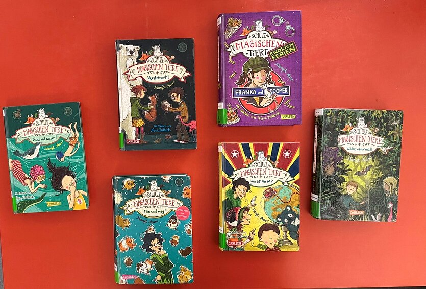 Farbige Fotos von Büchern der Reihe "Schule der magischen Tiere". Die Bücher liegen auf einem roten Tisch. - copyright: Stadtbibliothek Pforzheim - Ersteller: Stadtbibliothek Pforzheim