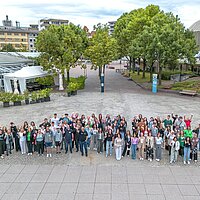 Viele Jugendliche auf einem Platz winken in die Kamera. Das Bild ist von oben aufgenommen.