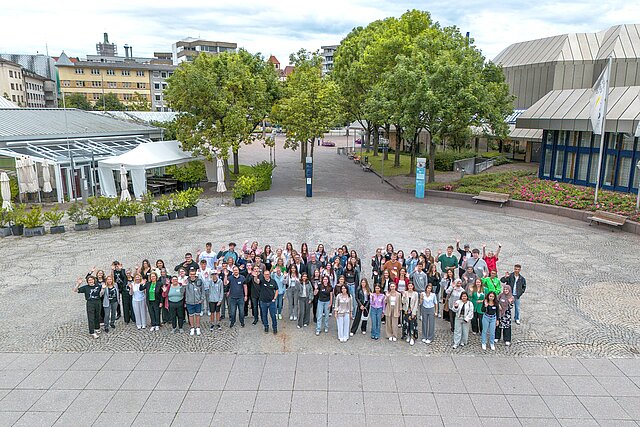 Viele Jugendliche auf einem Platz winken in die Kamera. Das Bild ist von oben aufgenommen.