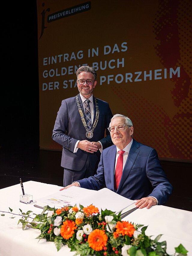 Ein Mann sitzt an einem Tisch, vor sich ein aufgeschlagenes Buch, ein Mann stehet hinter ihm. 