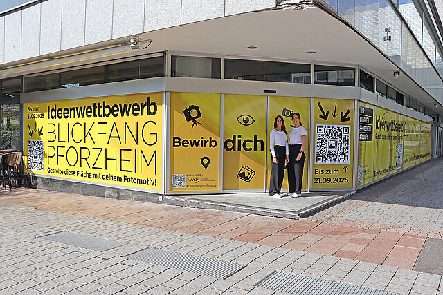 Zwei Frauen stehen vor einem mit gelber Folie beklebten Schaufenster mit der Aufschrift "Ideenwettbewerb Blickfang Pforzheim"