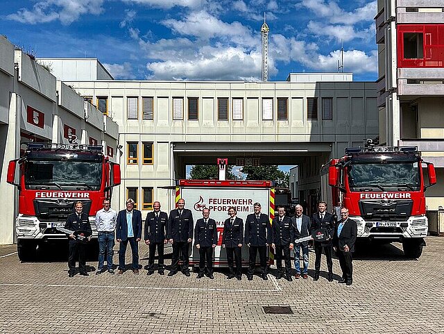 Farbfoto: Gruppenbild u. a. mit Feuerwehrkommandant Fischer  und Feuerwehr Fahrzeuge auf dem Gelände der Feuerwehr  