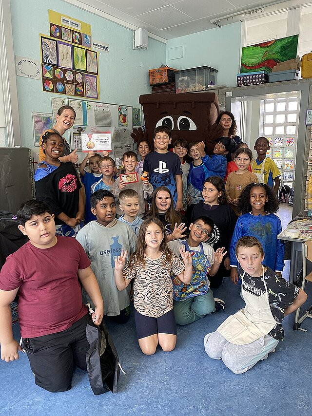 Auf dem Bild zu sehen sind Carina Aydin von der Firma UDATA und Tanja Leicht von der Abfallwirtschaft Pforzheim, die sich gemeinsam mit dem Maskottchen Bio-Tonni, das aussieht wie eine großé braune Bio-Tonne,  und Schülerinnen und Schüler der Insel Grundschule