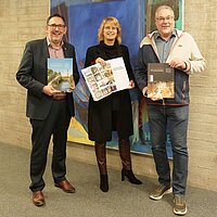 Drei Personen stehen vor einem Gemälde und halten je ein Magazin in den Händen.