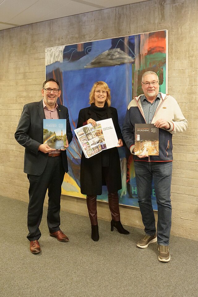 Drei Personen stehen vor einem Gemälde und halten je ein Magazin in den Händen.