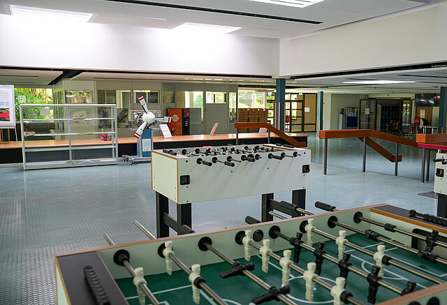 Eine Schulaula, die durch Deckenfenster hell beleuchtet ist. Im Vordergrund sind zwei Tischkicker zu sehen.