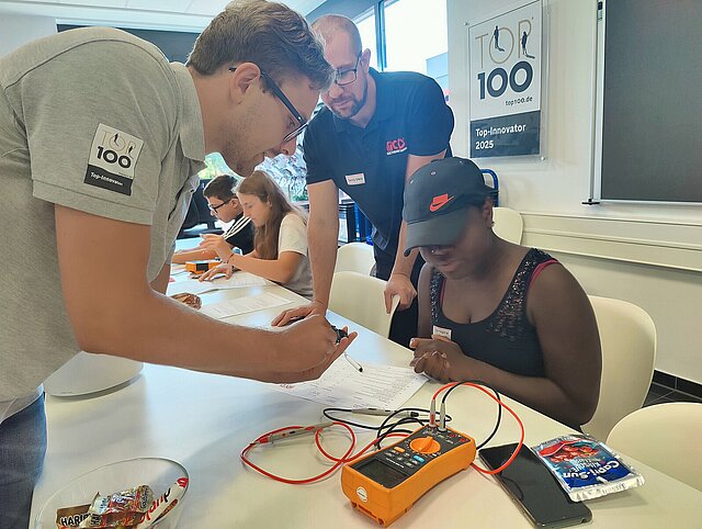 Mehrere Personen sitzen und stehen an einem Tisch und arbeiten gemeinsam an einem technischen Projekt mit einem Messgerät.