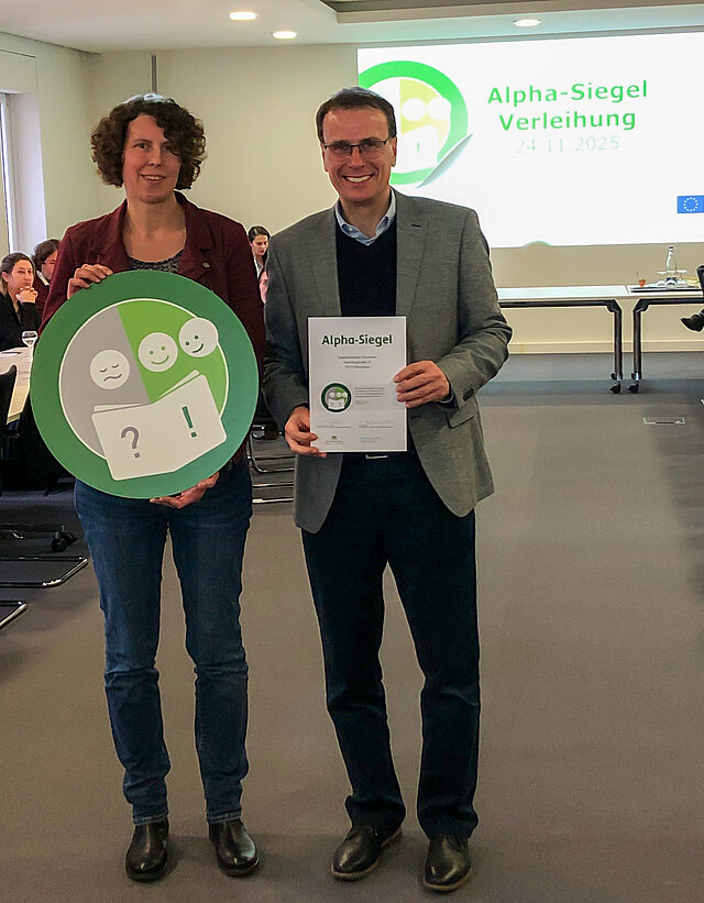 Auf dem Bild ist die tellvertretende Amtsleiterin der Stadtbibliothek, Marion Hekmann, zu sehen, die das Siegel aus der Hand von Staatssekretär Volker Schebesta, Ministerium für Kultus, Jugend und Sport, überreicht bekommt.