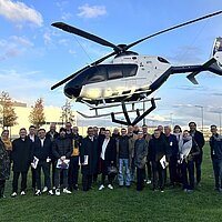 Eine Gruppe von Menschen steht vor einem in der Luft schwebenden Helikopter.