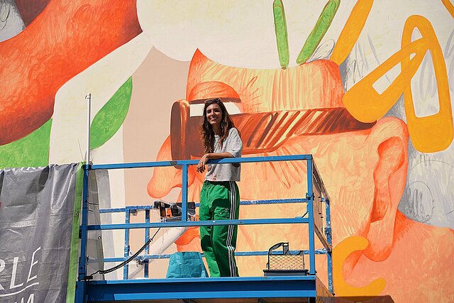 Künstlerin auf einem Scherenlift vor einer bunt bemalten Hausfassade