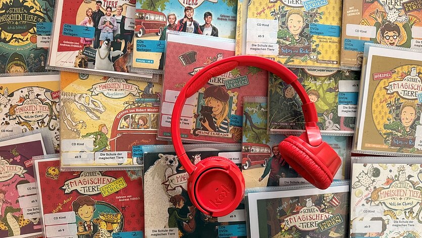 Ein roter Kinderkopfhörer liegt mittig auf vielen bunten CD-Hüllen der Hörspiel-Reihe „Die Schule der magischen Tiere“, mit gezeichneten Figuren und Tieren - copyright: Stadtbibliothek Pforzheim - Ersteller: Stadtbibliothek Pforzheim
