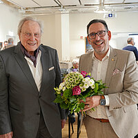 Zwei Männer im Anzug stehen vor vielen Menschen und schauen lächelnd in die Kamera. Ein Mann hält einen Blumenstrauß.