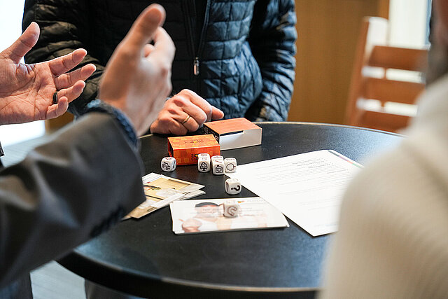 Ein Tisch, auf dem einige Würfel mit Symbolen darauf liegen. Im Vordergrund sind zwei gestikulierende Hände zu sehen.
