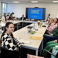 Mehrere Personen sitzen an Tischen in einer Fortbildungs-Situation, im Hintergrund ist ein eine Leinwand zu sehen.