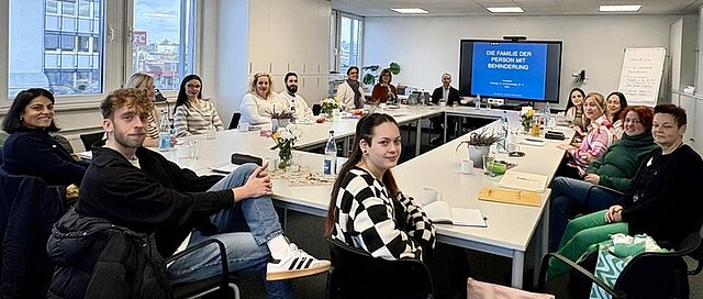 Mehrere Personen sitzen an Tischen in einer Fortbildungs-Situation, im Hintergrund ist ein eine Leinwand zu sehen.