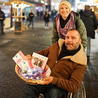 Ein Mann im Rollstuhl hält mit beiden Händen einen Korb, in dem leuchtende, lila Windlichter und Flyer sind. Hinter im steht eine Frau, die in die Kamera lächelt. Im Hintergrund sind beleuchtete Hütten und Besucher:innen eines Weihnachtsmarkts zu sehen. - copyright:miteinanderleben e.V.