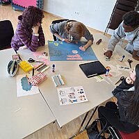 Auf dem Bild sind Jugendliche zu sehen, die um einen Tisch sitzen und stehen und ein Bild malen.