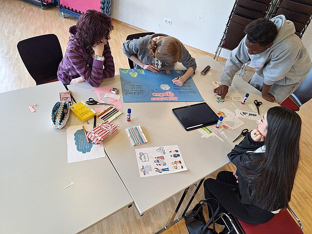 Auf dem Bild sind Jugendliche zu sehen, die um einen Tisch sitzen und stehen und ein Bild malen.