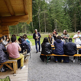 An zwei Tischen sitzen ungefähr 20 Personen, ein paar Personen stehen aus. Sie sind am Essen. Die Tische stehen vor einer Blockhütte am Waldrand. - copyright:Stadt Pforzheim