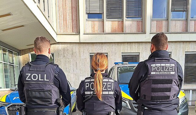 Farbfoto: stehend in einer Reihe rückwärts: Zollbeamter, Polizeibeamtin, Beamter der Polizeibehörde