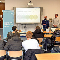 Ein Klassenzimmer, an einem Whiteboard stehen zwei Männer, die den Schülern etwas erklären.