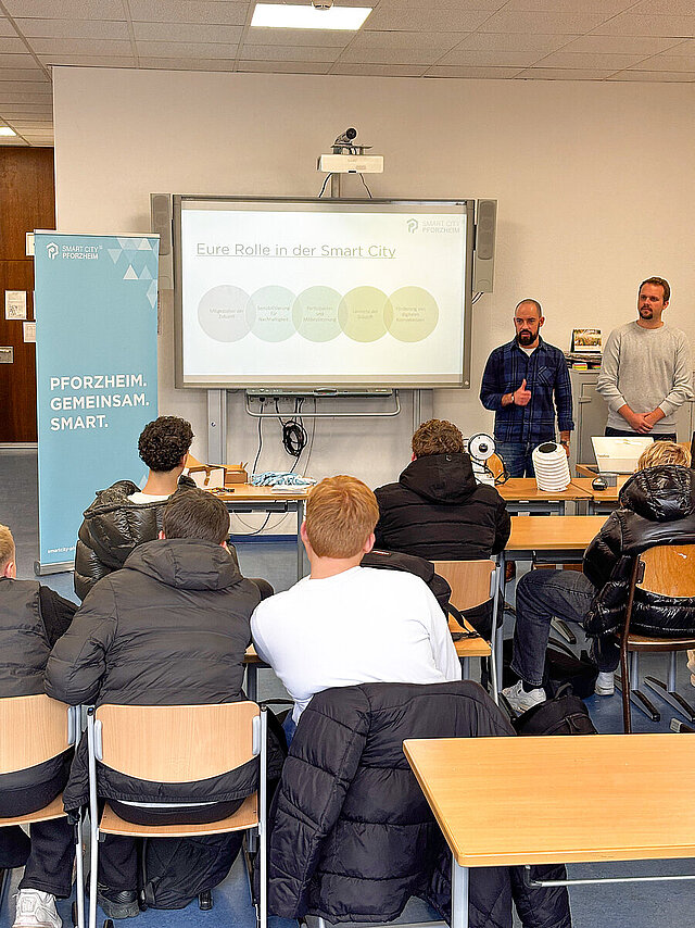 Ein Klassenzimmer, an einem Whiteboard stehen zwei Männer, die den Schülern etwas erklären.