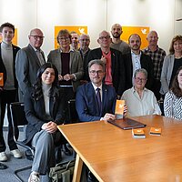 Mehrere Personen sitzen und stehen hinter einem Tisch und sehen in die Kamera