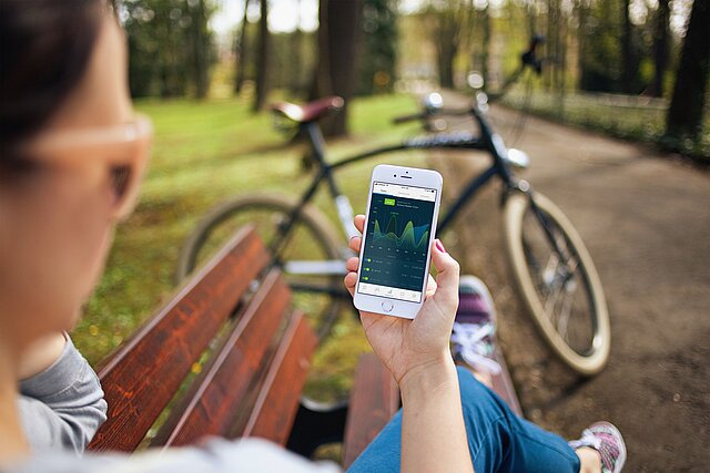 Frau hält Handy mit einer App auf dem Bildschirm in die Kamera. Im Hintergrund steht ein Fahrrad.