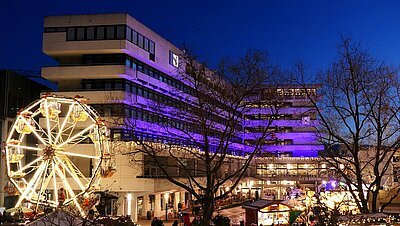 Auf dem Foto ist das Pforzheimer Rathaus zu sehen, das bei Dämmerung lila beleuchtet ist. Vor dem Rathaus auf dem Marktplatz ist der beleuchtete Weihnachtsmarkt zu sehen. Das Foto wurde von einer höheren Position aufgenommen.