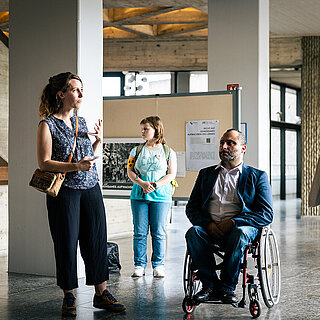 Drei Frauen und ein Mann im Rollstuhl hören einer sprechenden Frau zu. Rundherum sind Stellwänden zu sehen, an denen Portraits hängen. - copyright:Stadt Pforzheim