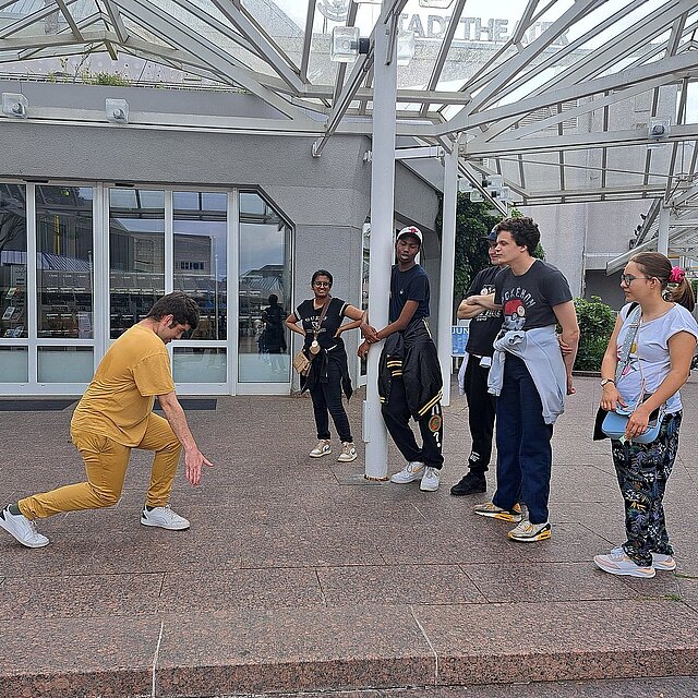 Ein junger Mann im gelben Outfit steht im Ausfallschritt vor einer Gruppe von fünf anderen Personen. - copyright:Stadt Pforzheim