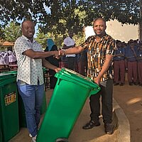 Zwei Männer aus Tansania im Vordergrund. Der eine übergibt eine grüne Mülltonne an den anderen Mann. Im Hintergrund sind Schüler zu sehen. Es handelt sich hierbei um die Umsetzung der Klimapartnerschaft der Stadt Pforzheim mit Mtwara in Tansania. 