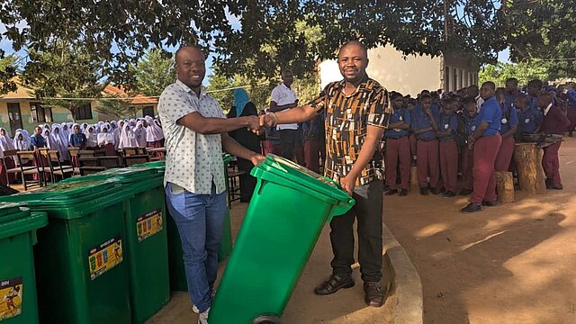 Zwei Männer aus Tansania im Vordergrund. Der eine übergibt eine grüne Mülltonne an den anderen Mann. Im Hintergrund sind Schüler zu sehen. Es handelt sich hierbei um die Umsetzung der Klimapartnerschaft der Stadt Pforzheim mit Mtwara in Tansania. 