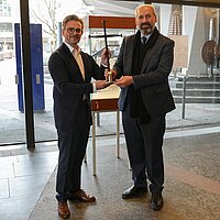 Zwei Männer im Anzug halten ein Kreuz in den Händen. Im Hintergrund ist eine Vitrine zu sehen.