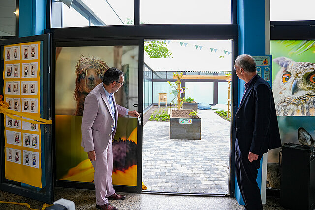 Zwei Männer im Anzug schauen durch eine offene Tür in einen neu gestalteten Innenhof. 