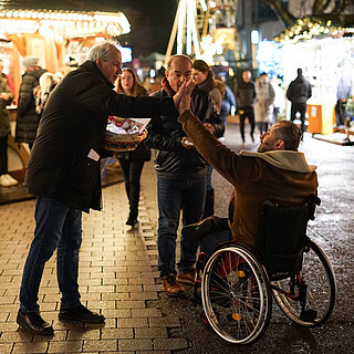 Ein großer Mann mit einen bunten Korb in der linken Hand gibt einem Mann im Rollstuhl ein "High Five".Im Hintergrund sind beleuchtete Hütten und Besucher:innen eines Weihnachtsmarkt zu sehen. - copyright:miteinanderleben e.V.