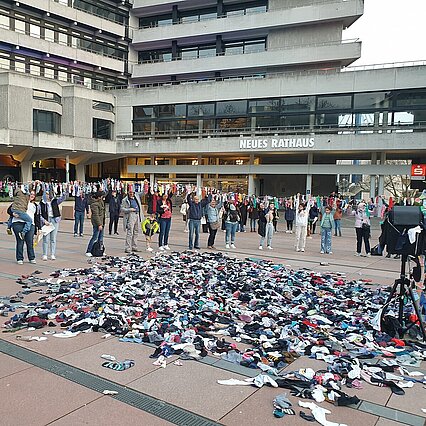 Im Vordergrund sieht man viele bunte Socken auf dem Boden liegen. Dahinter stehen Menschen, die eine Leine hoch halten. Auf der Leine sind Socken gehängt. Im Hintergrund sieht man das Rathaus Pforzheim. - copyright:Stadt Pforzheim