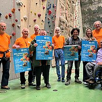 Die DAV Sektion Pforzheim, die Inklusionsbeauftragten der Stadt Pforzheim und des Enzkreises sowie der Verein Förderung Behinderter Pforzheim laden zum DAV-Erlebnistag 2026 ein. Die Mitglieder präsentieren das Plakat zur Veranstaltung vor einer Kletterwand.